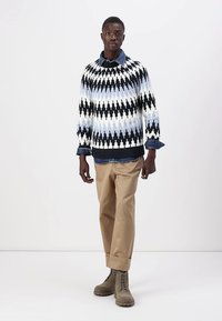 Maglione lavorato a maglia in un motivo geometrico nero, bianco e blu; pantaloni beige; camicia di denim visibile ai polsini, abbinata a stivali allacciati color caramello.