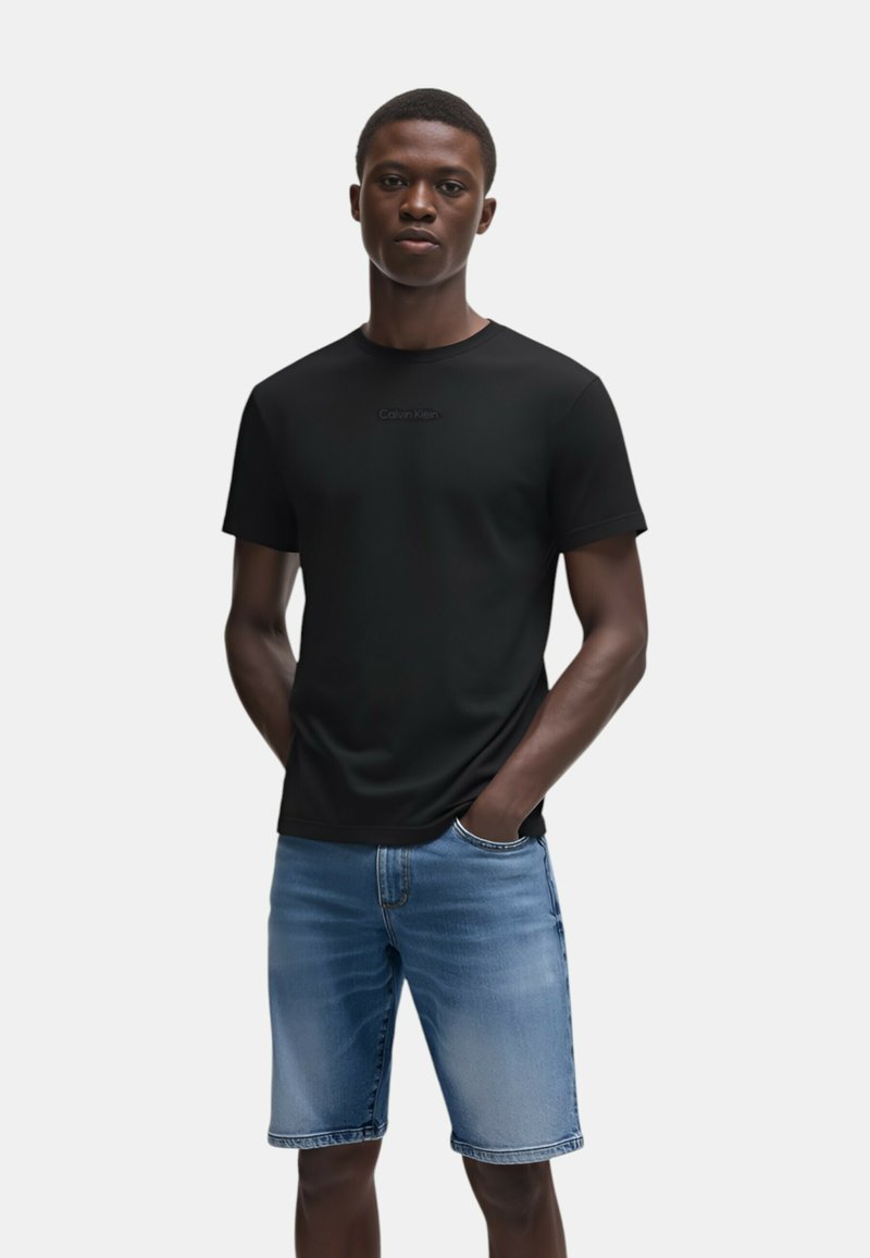 T-shirt nera a maniche corte con un logo Calvin Klein discreto, abbinata a pantaloncini di jeans azzurri chiari. Il tessuto appare liscio e informale.