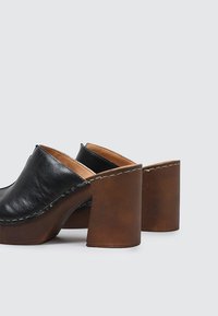Zuecos negros de piel sin cierre, con tacones cuadrados de madera marrón y costuras visibles a lo largo del borde superior, mostrados sobre un fondo liso.
