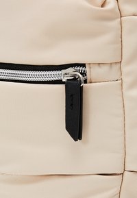 Gros plan sur un tissu beige avec une fermeture éclair argentée et une languette en cuir noir sur un sac ou une veste matelassée.