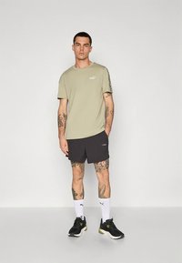 Olive green t-shirt with logo, paired with black shorts. Model wears black sneakers and white crew socks, showcasing detailed tattoos on arms.