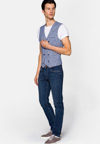 Niebieska i biała kamizelka w kratę na białym T-shircie, zestawiona z ciemnymi dżinsami i szarymi sneakersami. Posiada podwójne guziki.