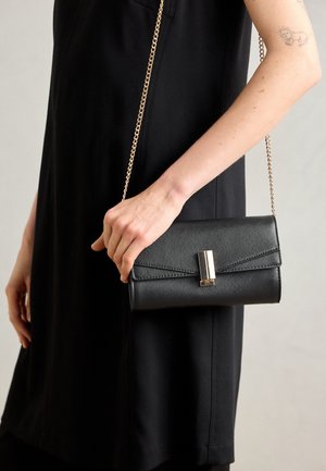 Black leather crossbody bag with a textured finish, gold hardware, flap closure, and a chain strap, held by a person in a dark outfit.