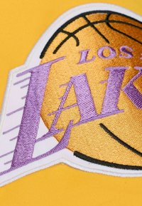 Logo der Los Angeles Lakers mit lila und goldener Stickerei auf gelbem Hintergrund, einschließlich einer Basketballgrafik und einem stilisierten Textdesign.