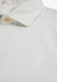 Camisa blanca de algodón con acabado texturizado, que presenta un pequeño logo bordado de una calavera blanca en el pecho.