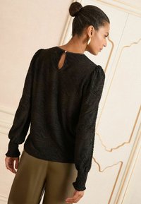 Blusa nera in tessuto strutturato con maniche lunghe a sbuffo, caratterizzata da un dettaglio a chiave sul retro e un motivo ondulato. Abbinata a pantaloni verde oliva.
