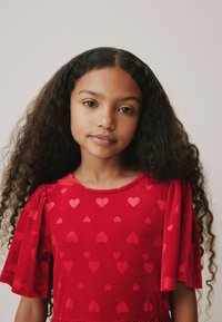 Vestito rosso con motivo a cuori, maniche corte a sbuffo e scollo rotondo. Tessuto morbido con una leggera lucentezza. Finitura del tessuto liscia in tutta la superficie.