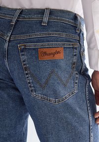 Közeli kép egy kék Wrangler farmer hátul zsebéről, barna varrott „W” díszítéssel és bőr márkajelzéssel a zseb felett, fehér inggel viselve.