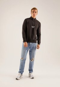 Fekete cipzáras pulóver "BOSTON" felirattal, világoskék szakadt farmernadrággal és szürke tornacipővel párosítva. Sima anyag, laza szabás, minimális részletekkel.