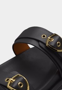 Μαύρο δερμάτινο σανδάλι με δύο ρυθμιζόμενους ιμάντες που διαθέτουν αγκράφες σε χρυσό τόνο, σε απλό φόντο.