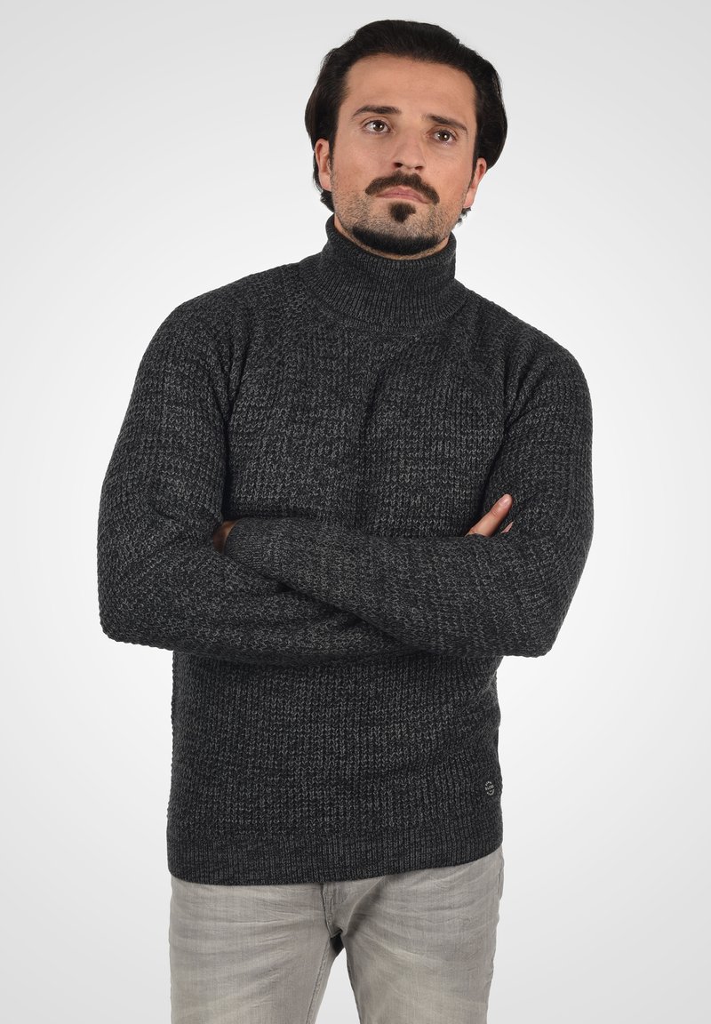 Pull en col roulé gris foncé avec un tricot texturé, présentant des poignets et un ourlet côtelés, porté avec un pantalon gris clair.