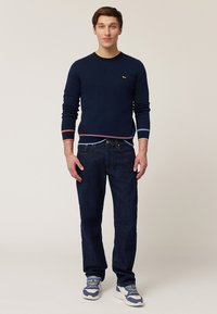 Marineblauer Rundhals-Pullover mit gelbem Kragen, roten und blauen Streifen an den Bündchen und dem Saum, kombiniert mit dunkelblauen Jeans und weißen Sneakers.
