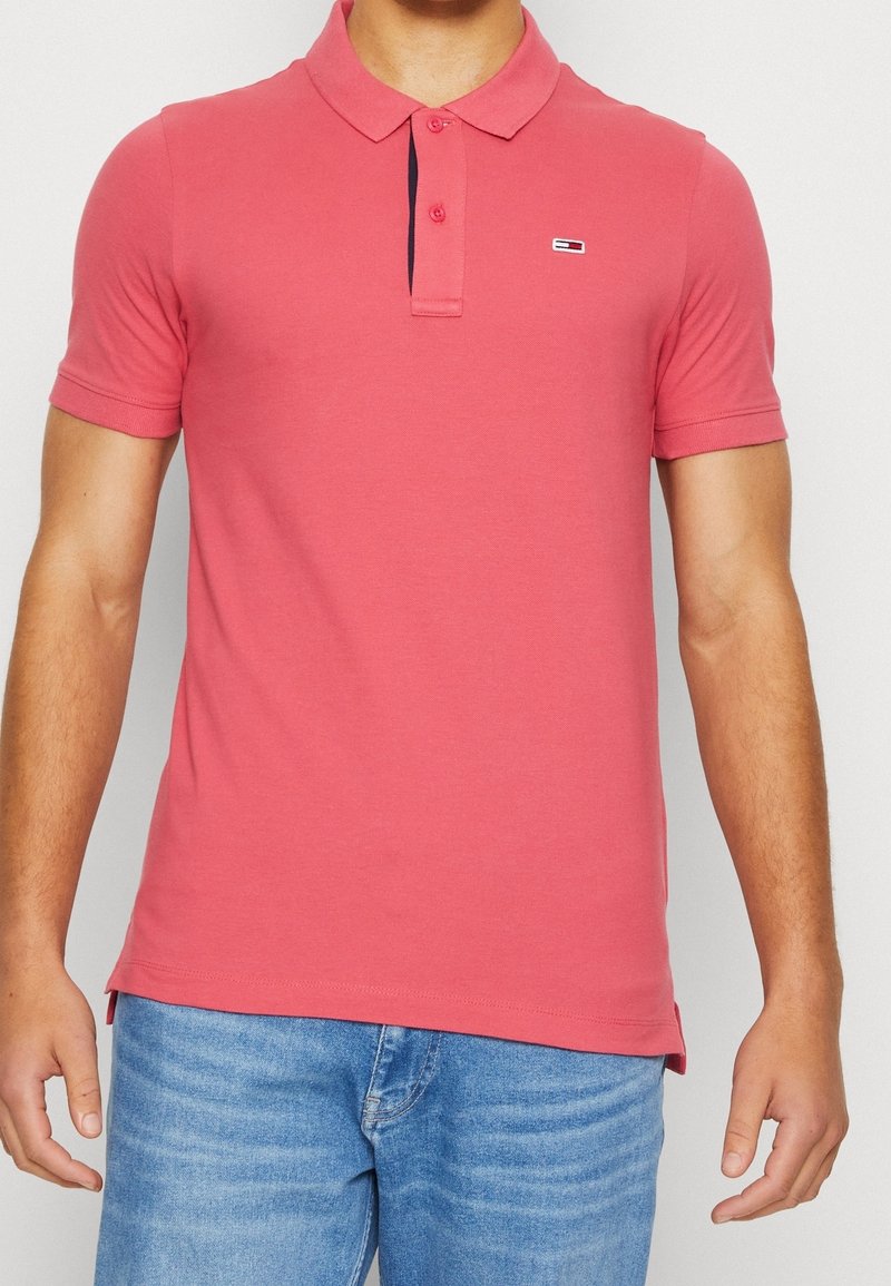 Homme portant un polo rose corail ajusté à manches courtes avec deux boutons et un petit logo sur la poitrine gauche, associé à un jean bleu clair.