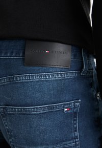 Modré denimové džínsy s čiernym koženým pásom, na ktorom je odtlačené "TOMMY HILFIGER." Obsahujú kontrastný steh a malý akcent loga.