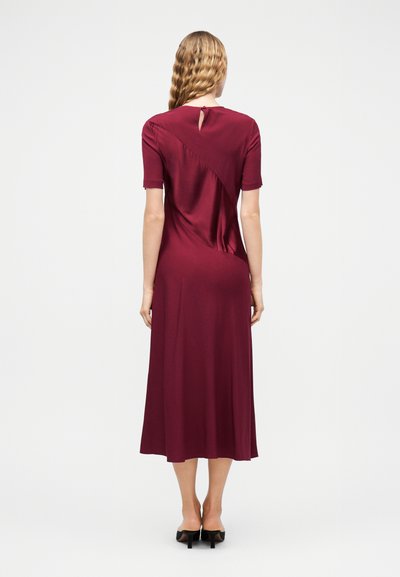 Γυναίκα με κυματιστά ξανθά μαλλιά που φοράει μπέρντιλαν επιμήκους μήκους, με κοντά μανίκια, μπορντό φόρεμα με άνοιγμα στην πλάτη και μαύρα ψηλοτάκουνα παπούτσια.