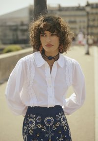 Femme aux cheveux bouclés portant une blouse blanche à volants et une jupe bleue avec broderies florales, debout en plein air près d'un poteau dans un cadre urbain.