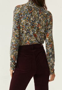 Donna che indossa una blusa a maniche lunghe con motivo floreale infilata in pantaloni di velluto a coste viola scuro a vita alta, mostrata di spalle.