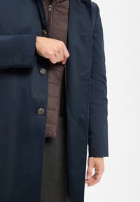 Man in een marineblauwe jas die over een bruine gewatteerde jas is geknoopt, terwijl hij zich aan het zippen is, wat gelaagde winterkleding toont op een witte achtergrond.