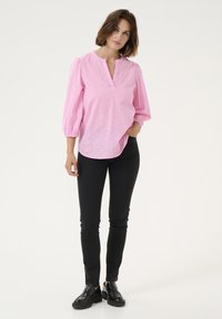 Blouse rose avec des manches bouffantes, décolleté en V et motif texturé subtil. Associée à un pantalon noir slim et des mocassins noirs.