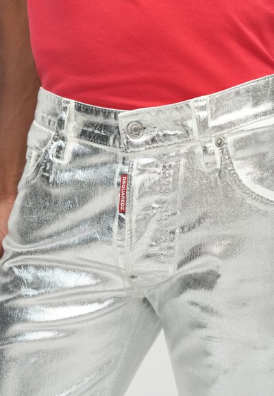 Hõbedased metallik teksad läikiva tekstuuriga, millel on ees nööpidega kinnitamine, taskud ja nähtav brändimärgistus vööl.