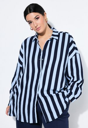 Kobieta o ciemnych włosach nosi oversize koszulę z pionowymi jasnoniebieskimi i ciemnoniebieskimi paskami oraz ciemne spodnie, stoi na białym tle.