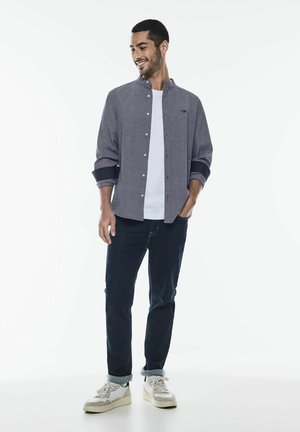 Camisa de botones gris con cuello mao, mangas enrolladas, superpuesta a una camiseta blanca, combinada con jeans oscuros y zapatillas blancas.