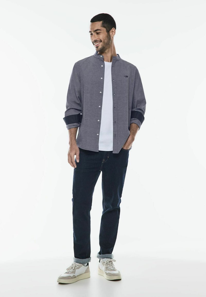 Camicia grigia con colletto alla coreana, maniche arrotolate, sovrapposta a una t-shirt bianca, abbinata a jeans scuri e sneaker bianche.