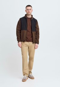 Brun og svart fleecejakke med en glidelåslomme, brun skjorte, khaki bukser og grå joggesko med hvite lisser. Enkelt design, uformelt utseende.
