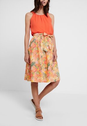 Femme portant un haut orange sans manches avec une jupe fleurie longueur genou et des sandales beige, debout devant un fond uni.