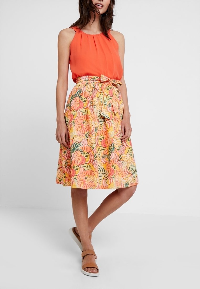 Femme portant un haut orange sans manches avec une jupe fleurie longueur genou et des sandales beige, debout devant un fond uni.