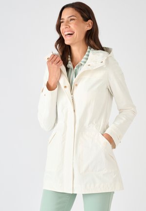 Femme souriante portant une veste à capuche beige clair sur une chemise rayée verte et blanche, avec une main dans la poche de la veste.