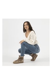 Pull en maille épaisse beige avec un col en V, jeans bleus ajustés et bottes mi-mollet marron avec une finition texturée et des lacets.