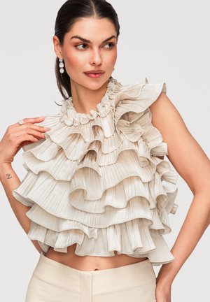 Femme portant un haut beige sans manches à volants avec des plis superposés, regardant sur le côté, les cheveux tirés en arrière et portant des boucles d'oreilles pendantes.