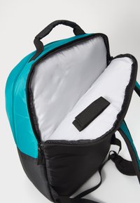 Rucksack mit türkisfarbenen und schwarzen Abschnitten, ausgestattet mit einem Reißverschluss, grauer Innenauskleidung und einem Klettverschlussriemen zur sicheren Befestigung von Gegenständen im Inneren.