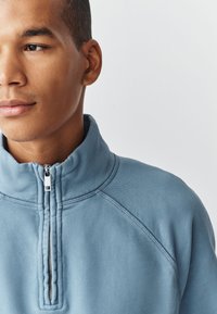 Noor mees kandes helesinist kvartslukuga dressipluusi, vaadates veidi vasakule ühevärvilise valge tausta ees.