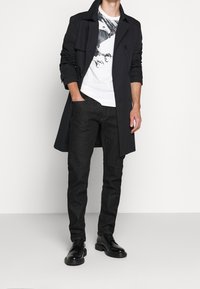 Černý kabát přes bílý tričko s grafickým potiskem zobrazujícím ptáka. Tmavé džíny a černé boty, vytvářející vícevrstvý outfit.