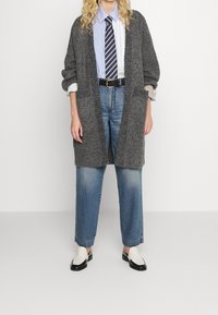 Grå stickad kofta med öppen framkant, bärs över en blåvit randig skjorta, kombinerad med ljusblå jeans och tvåfärgade loafers.