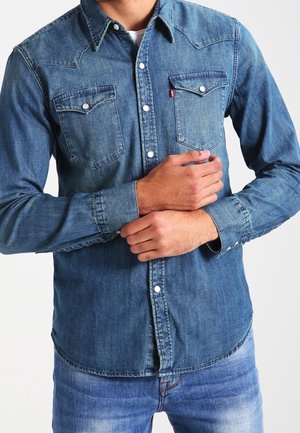 Homme portant une chemise en denim bleu à manches longues avec des boutons-pression et un jean bleu, tenant un poignet avec la main opposée.