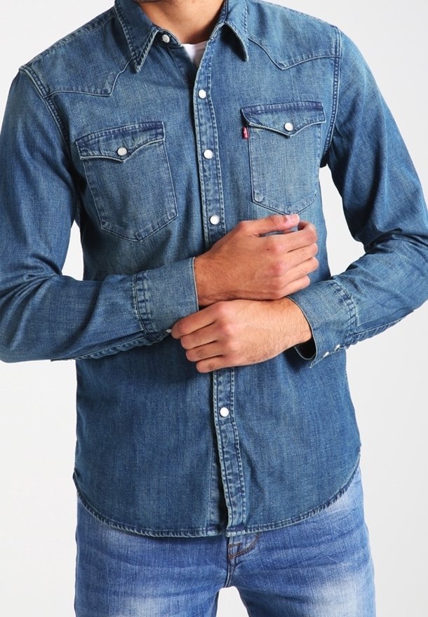 Homme portant une chemise en denim bleu à manches longues avec des boutons-pression et un jean bleu, tenant un poignet avec la main opposée.