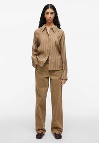 Donna con capelli lunghi e scuri, che indossa una camicia beige a righe con bottoni, pantaloni marroni larghi e scarpe basse marroni scure, in piedi su uno sfondo bianco.