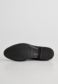Suela de zapato negra con goma texturizada, patrón en zigzag, bordes suaves y una impresión de logo circular.
