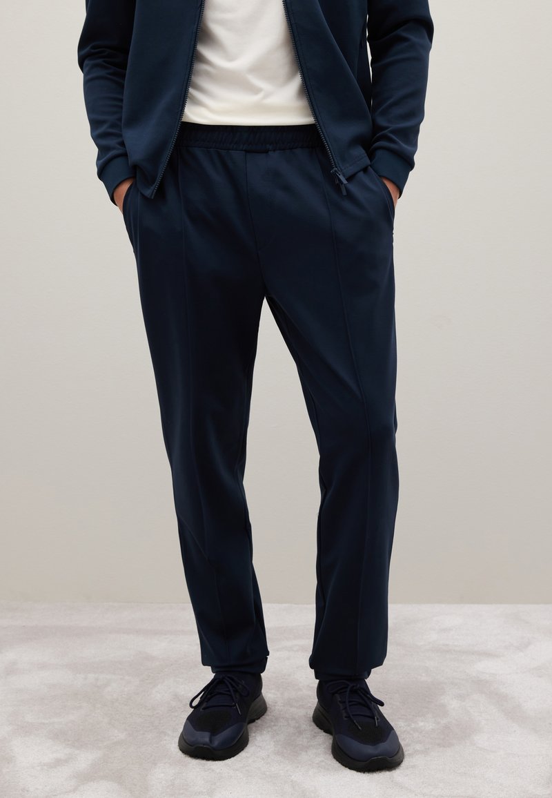 Pantaloni sportivi blu navy con vita elasticizzata e tasche laterali, realizzati in tessuto liscio e elasticizzato, abbinati a scarpe sportive nere.