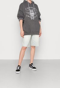 Osoba ubrana w ciemnoszary hoodie z grafiką czaszki, jasnoniebieskie szorty do kolan oraz czarne trampki niskie z białymi sznurowadłami.