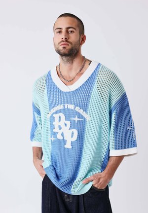 Hombre con pelo corto y barba que lleva un suéter de malla azul claro y blanco con el texto "Change the Game", de pie con las manos en los bolsillos.