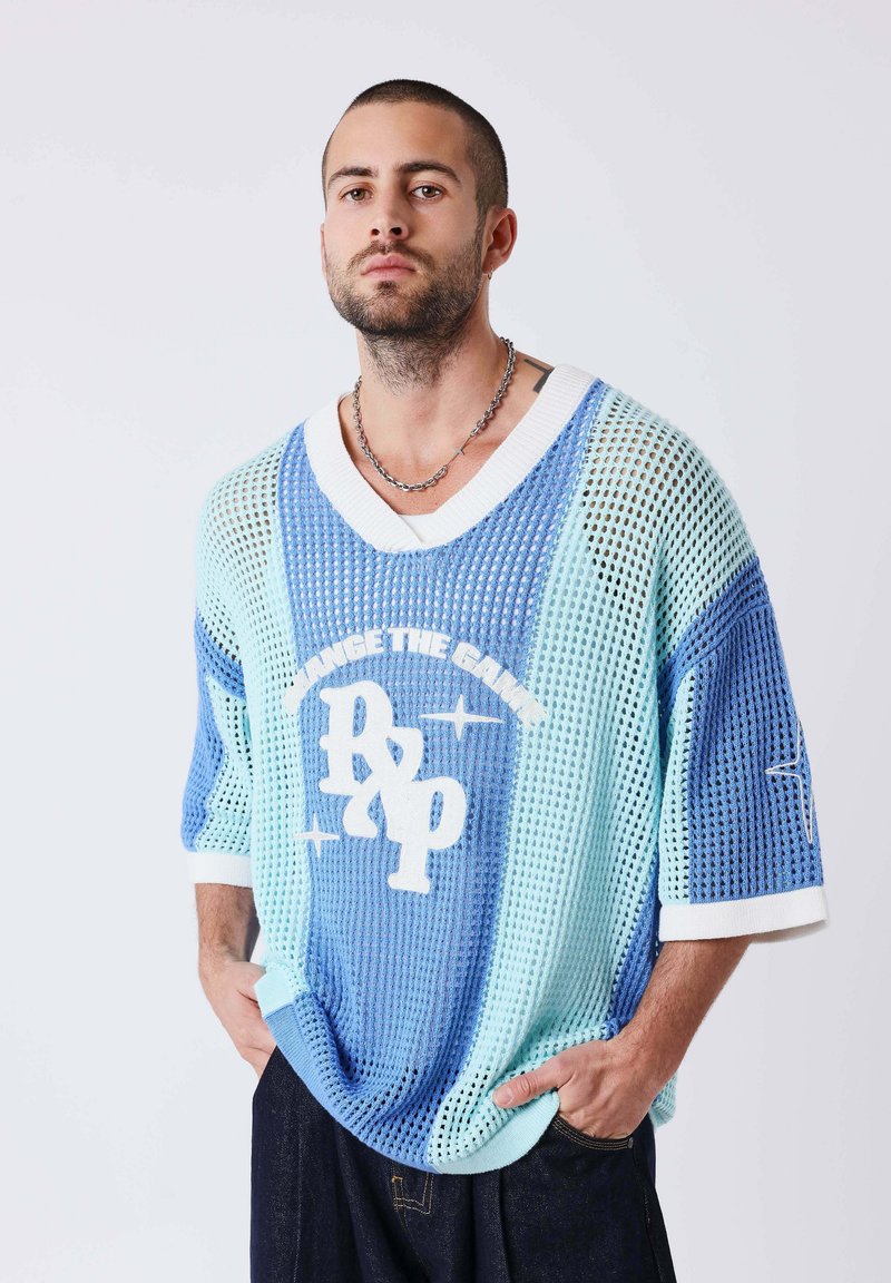 Homme aux cheveux courts et à la barbe, portant un pull en maille bleu clair et blanc avec l'inscription « Change the Game », debout les mains dans les poches.