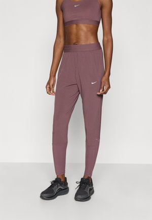 Persona atlética vistiendo un sujetador deportivo Nike malva y pantalones joggers a juego con zapatos de correr negros, de pie contra un fondo claro y liso.