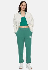Valkoinen bomber- takki vihreillä raidoilla, vihreä crop-top ja teal- väriset verkkarit "Fila Sports" -logolla, yhdistettynä valkoisiin lenkkareihin.