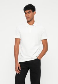 Camisa polo branca com colarinho, com abertura de dois botões e mangas curtas. Usada com calças pretas, apresentando um design slim fit e textura suave.
