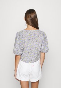 Femme avec de longs cheveux bruns portant des boucles d'oreilles créoles, un blouse à manches courtes à motifs floraux, et un short blanc retroussé, se tenant de dos par rapport à la caméra.