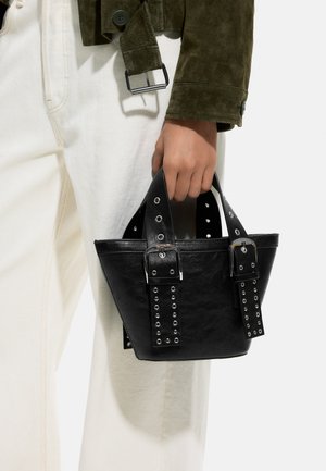 Ręka trzyma małą czarną skórzaną torebkę z podwójnymi paskami na klamry, w ubraniu oliwkowo zielonej kurtce i jasnych spodniach.